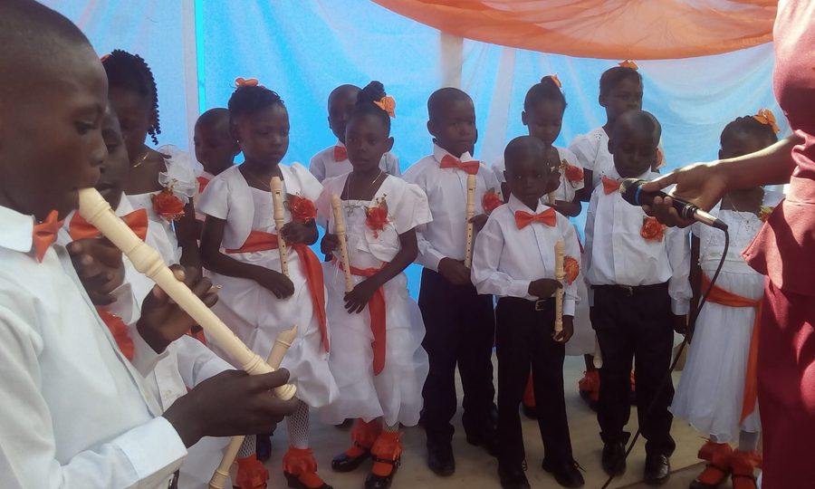 A graduate playing the recorder into a microphone while classmates look on