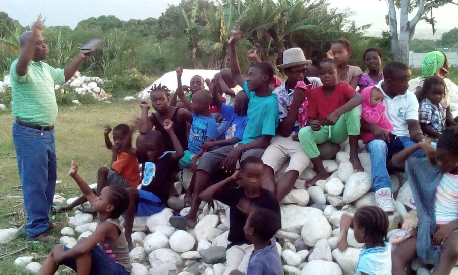 Children sitting outdoors with hands raised during a children&rsquo;s ministry gathering